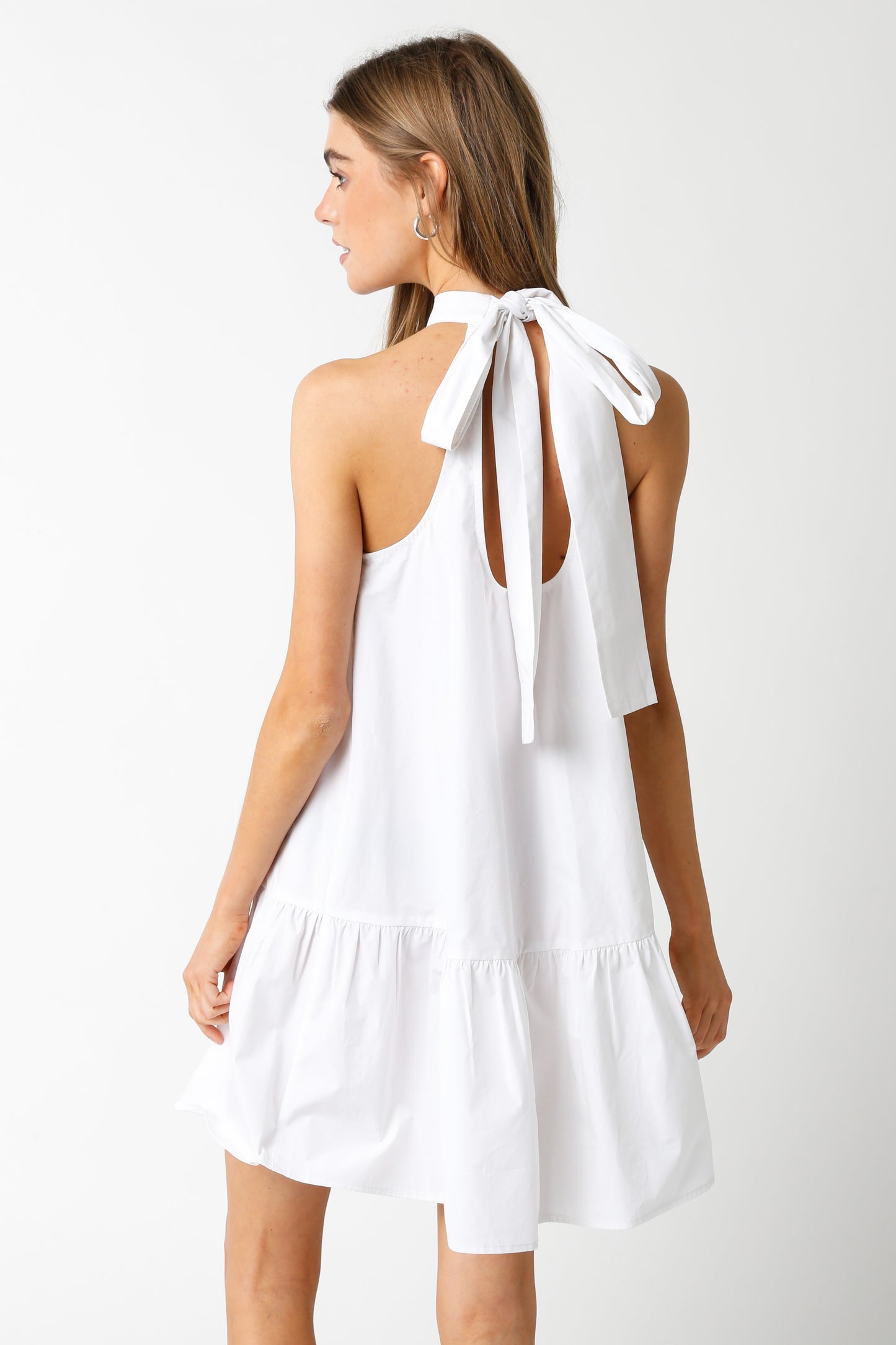 Woman wearing a sleeveless white halter mini dress with a high neckline and a bow-tie back detail.