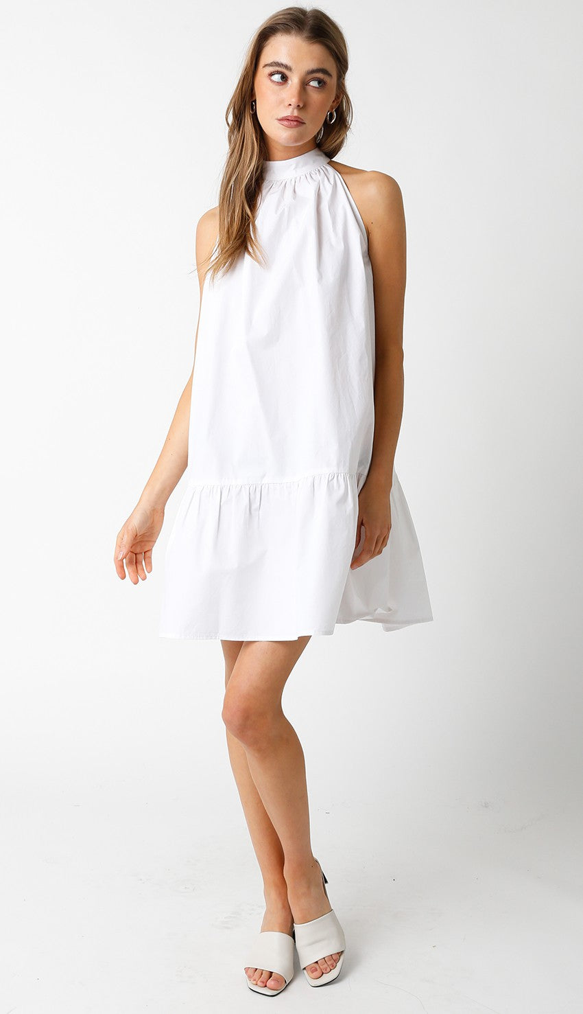 Woman wearing a sleeveless white halter mini dress with a high neckline and a bow-tie back detail.