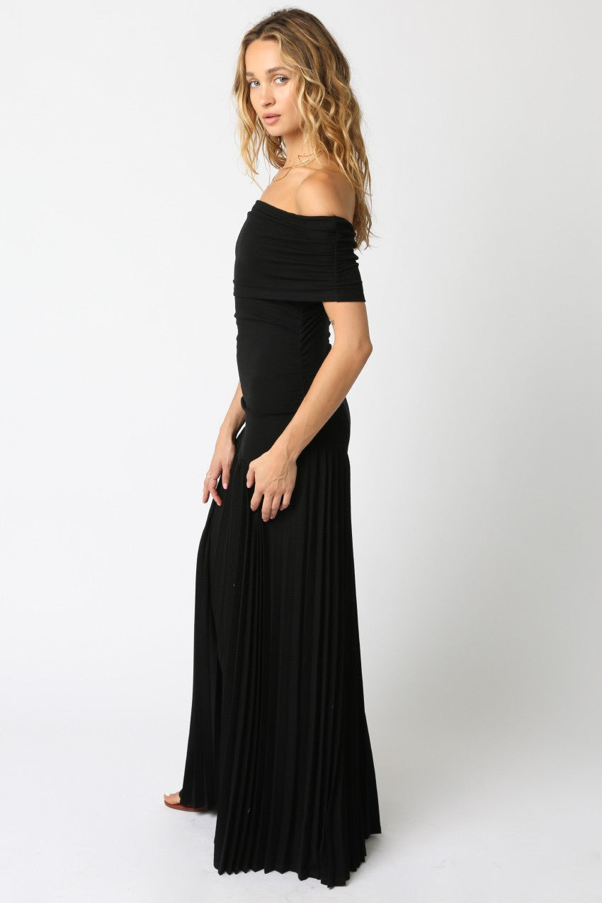 side view of Woman wearing a black off-the-shoulder maxi dress with a fitted bodice and pleated floor-length skirt, standing against a white background.