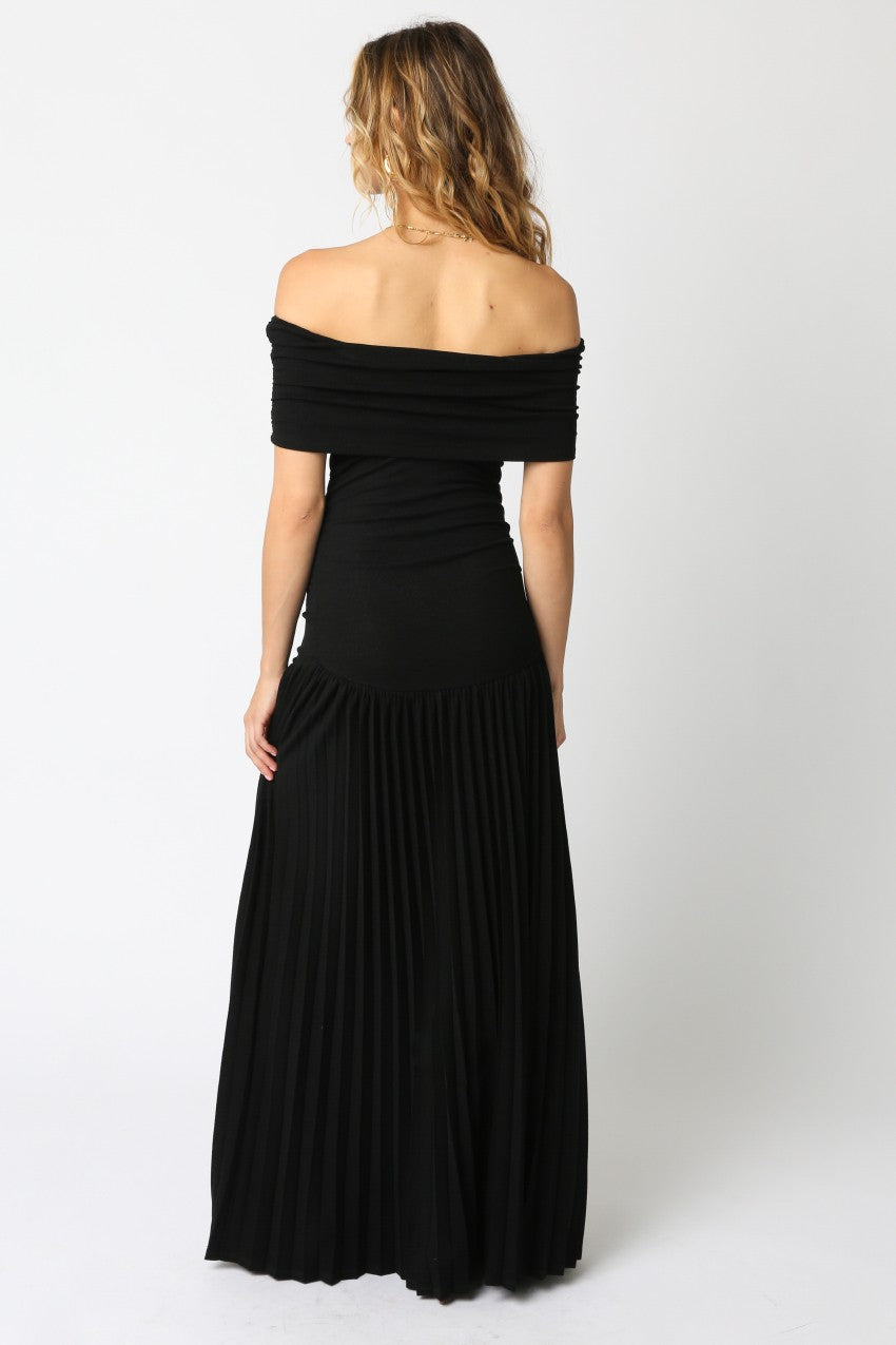 back view Woman wearing a black off-the-shoulder maxi dress with a fitted bodice and pleated floor-length skirt, standing against a white background.