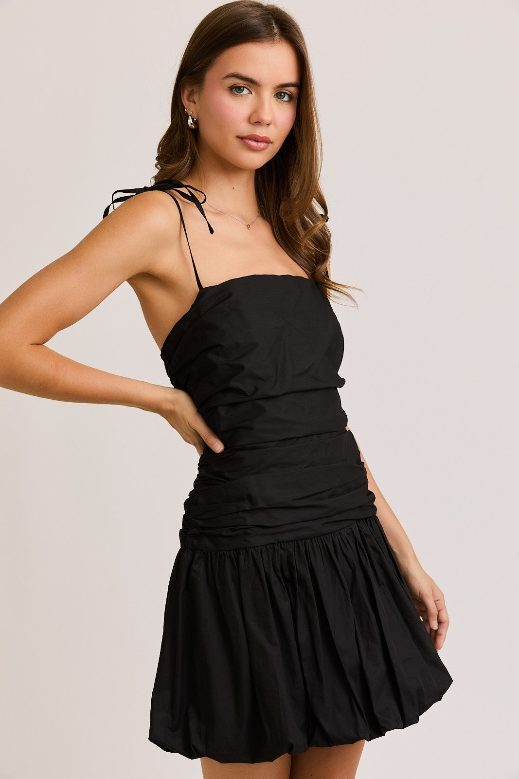 Woman wearing a black ruched mini dress with adjustable tie straps and a bubble hem, creating a chic and flirty silhouette perfect for a night out or special occasion.