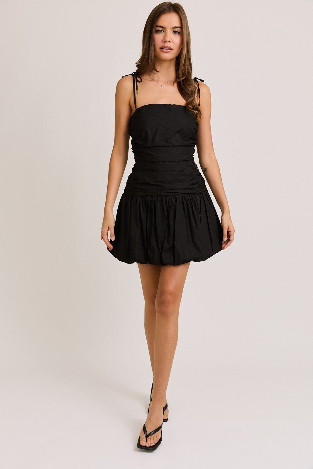Woman wearing a black ruched mini dress with adjustable tie straps and a bubble hem, creating a chic and flirty silhouette perfect for a night out or special occasion.