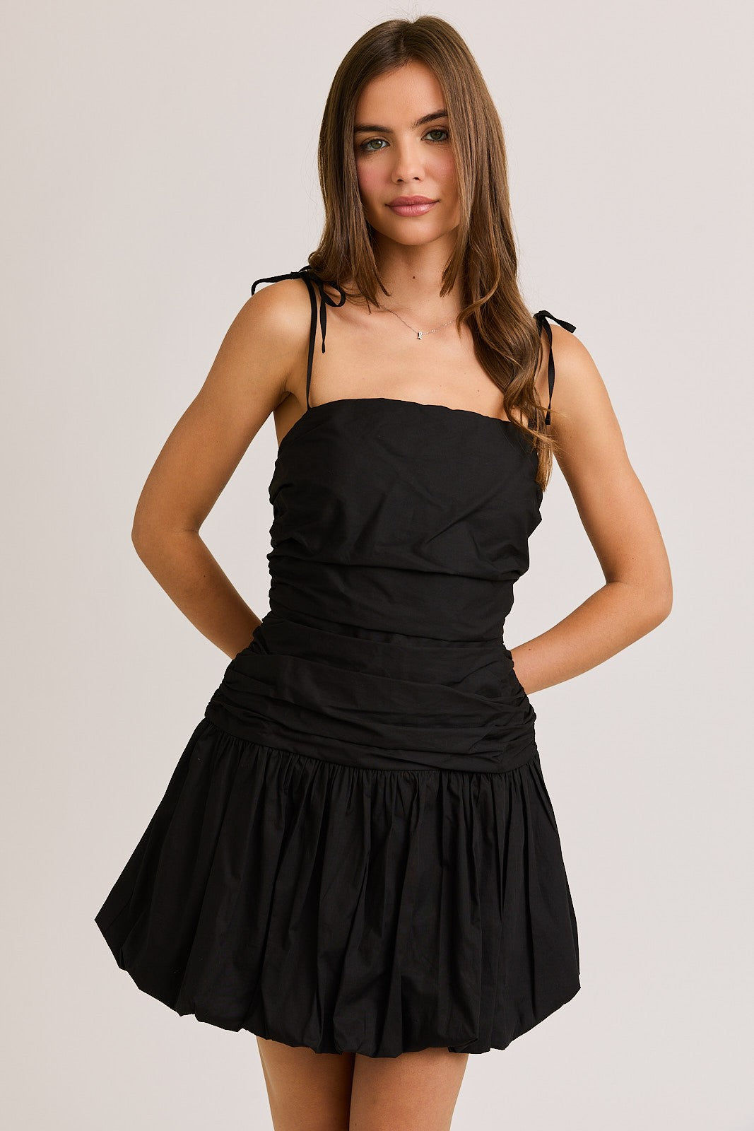 Woman wearing a black ruched mini dress with adjustable tie straps and a bubble hem, creating a chic and flirty silhouette perfect for a night out or special occasion.