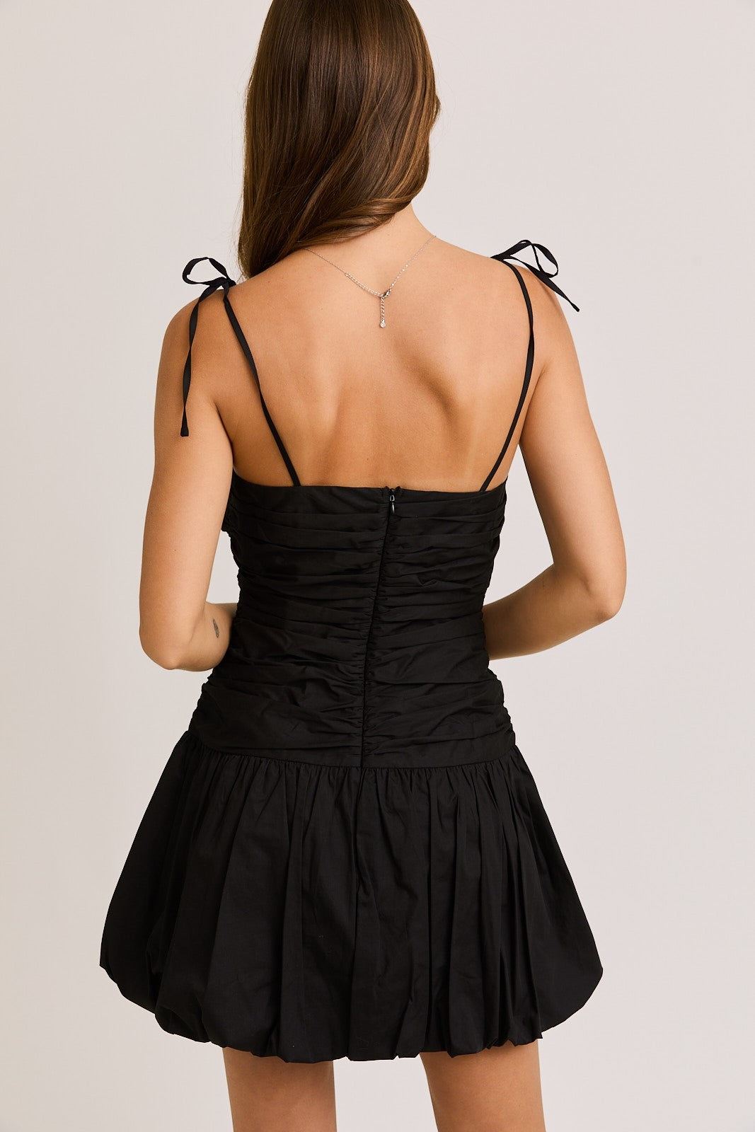 Woman wearing a black ruched mini dress with adjustable tie straps and a bubble hem, creating a chic and flirty silhouette perfect for a night out or special occasion.
