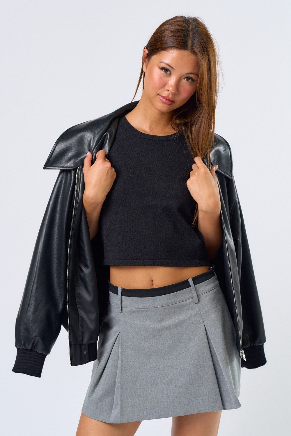 Woman wearing a black leather jacket, black crop top, and gray pleated skirt on a white background