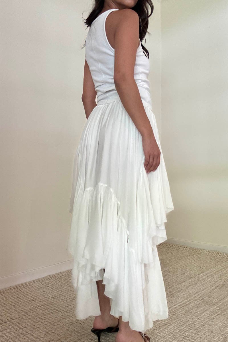 Back view of a woman in a white tank top and a long, asymmetrical white ruffle skirt, standing barefoot on a textured carpet.