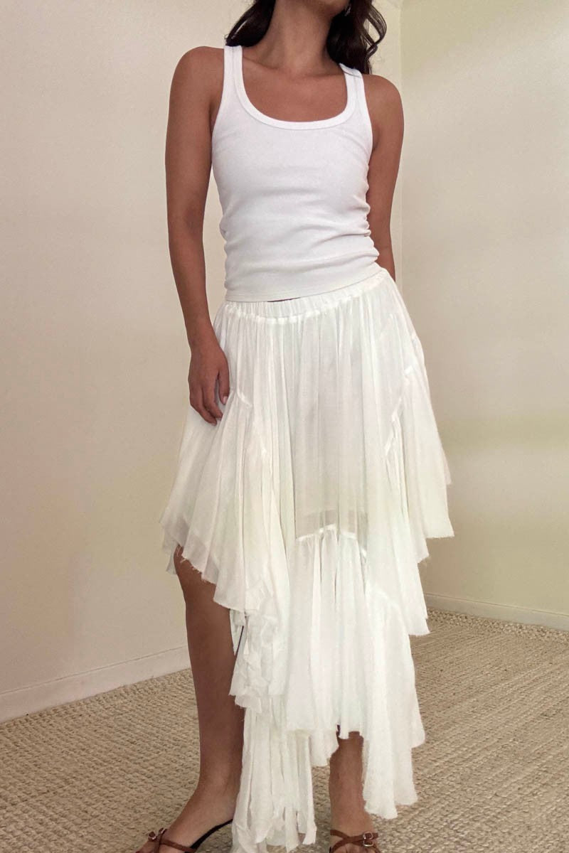 Woman wearing a white tank top and an asymmetrical white ruffle midi skirt, standing in a neutral-toned room with a textured carpet.