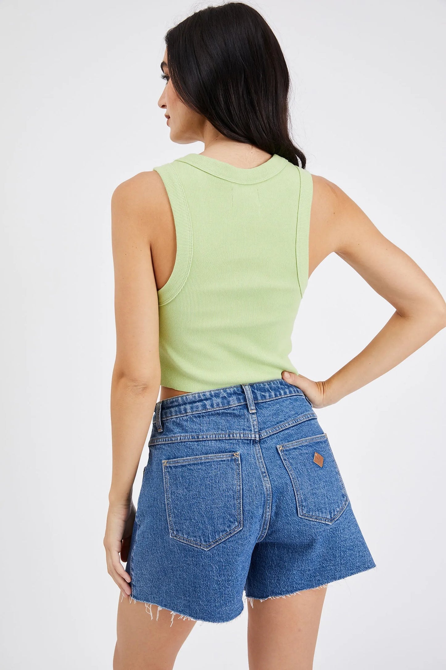 back view Woman wearing high-waisted, mid-length denim shorts with a raw hem and classic five-pocket styling, paired with a green crop tank top and black platform sandals.