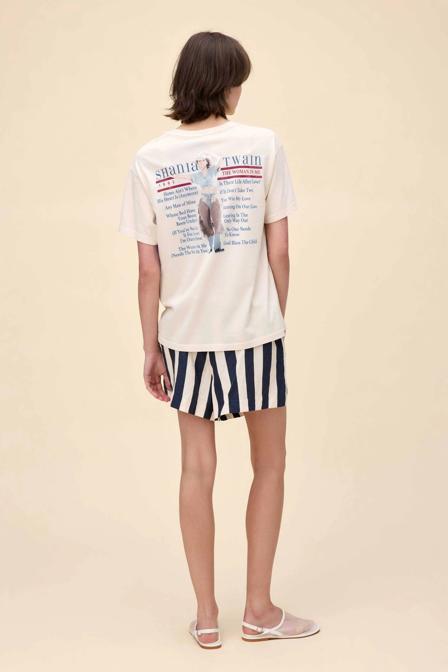 Back view of a cream-colored Shania Twain graphic tee featuring a 1995 “The Woman in Me” tour-inspired design with a photo of Shania in western attire and a list of song titles printed in red and blue text.