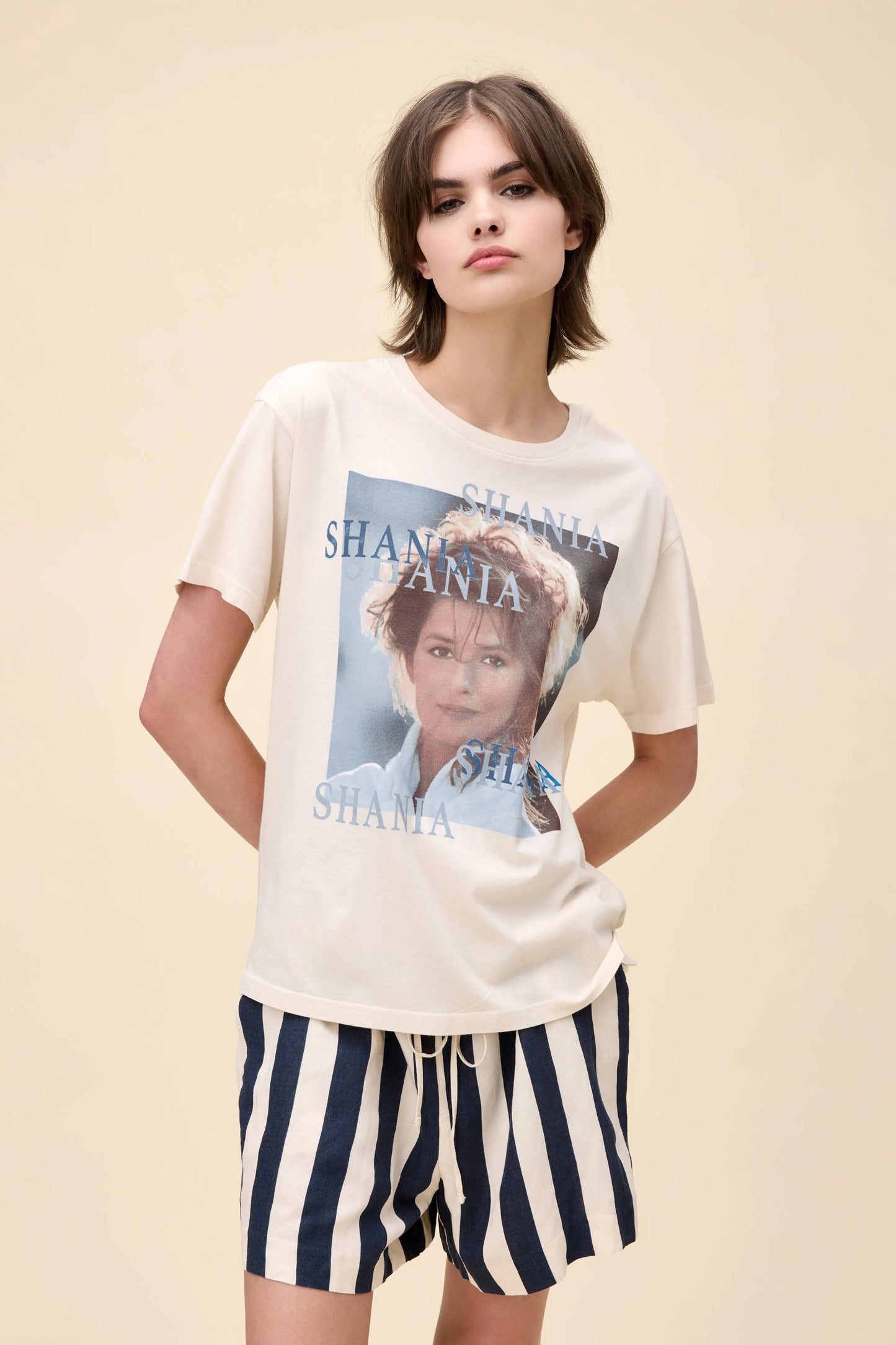 Woman wearing a cream-colored Shania Twain graphic tee with multiple "Shania" text overlays and a portrait-style image on the front, paired with navy and white striped shorts.
