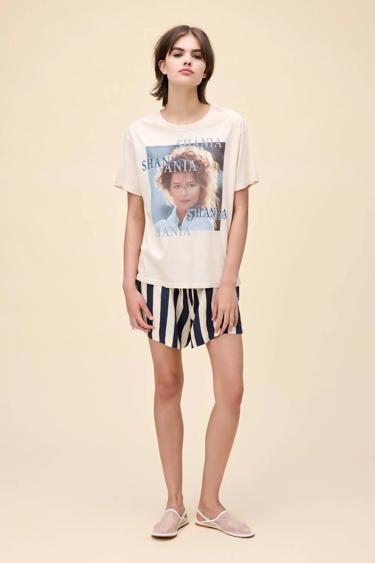 Woman wearing a cream-colored Shania Twain graphic tee with multiple "Shania" text overlays and a portrait-style image on the front, paired with navy and white striped shorts.