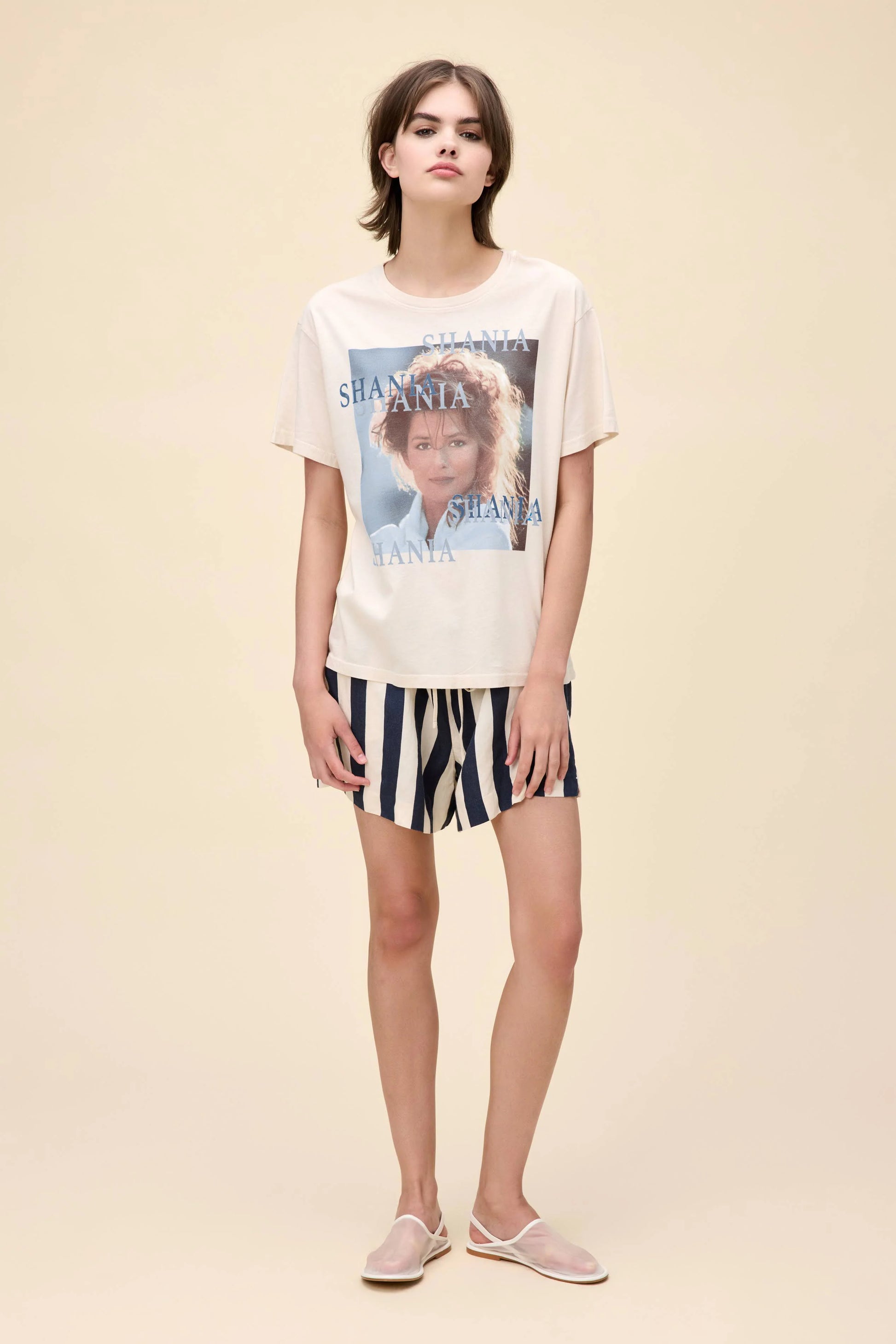 Woman wearing a cream-colored Shania Twain graphic tee with multiple "Shania" text overlays and a portrait-style image on the front, paired with navy and white striped shorts.
