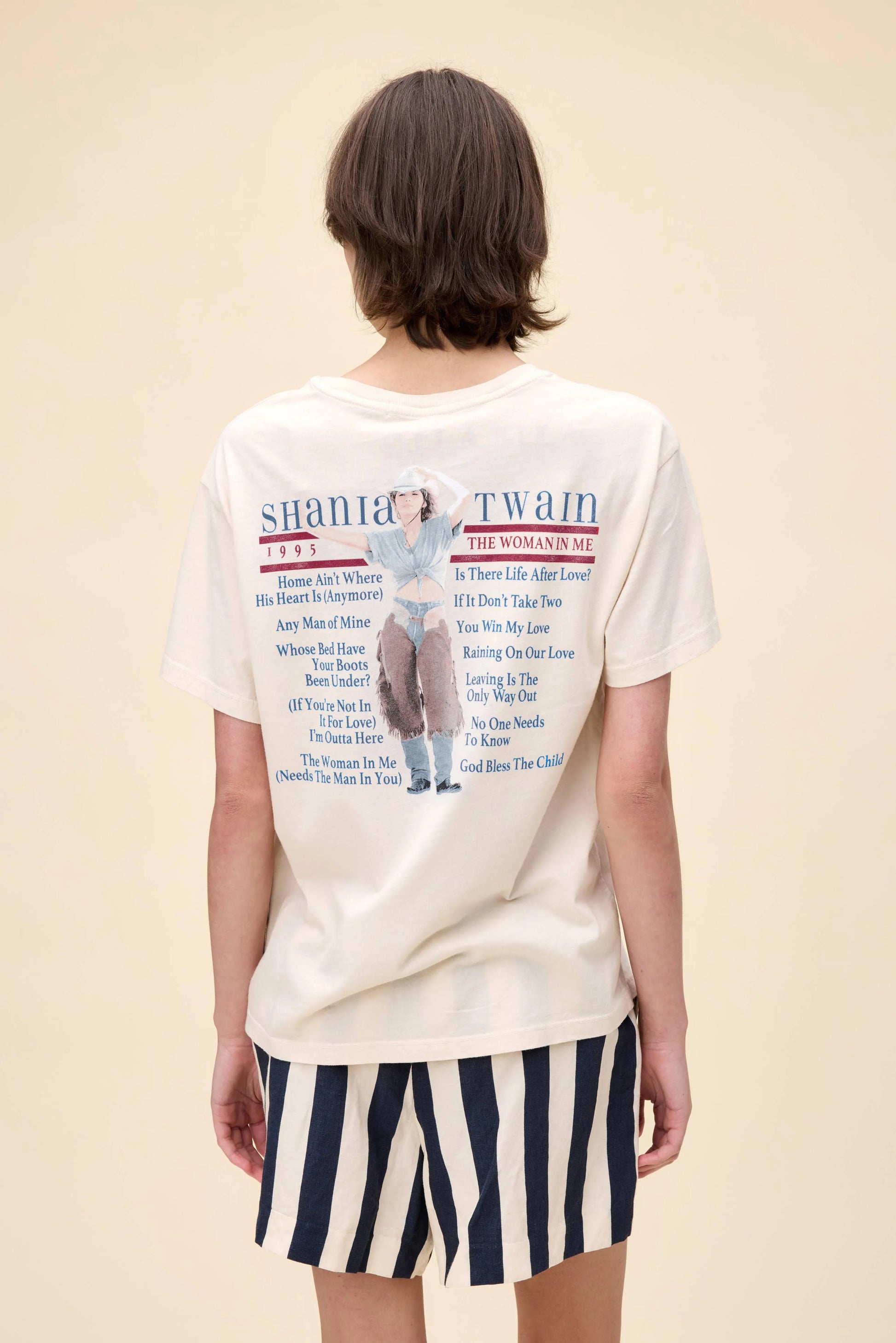 Back view of a cream-colored Shania Twain graphic tee featuring a 1995 “The Woman in Me” tour-inspired design with a photo of Shania in western attire and a list of song titles printed in red and blue text.
