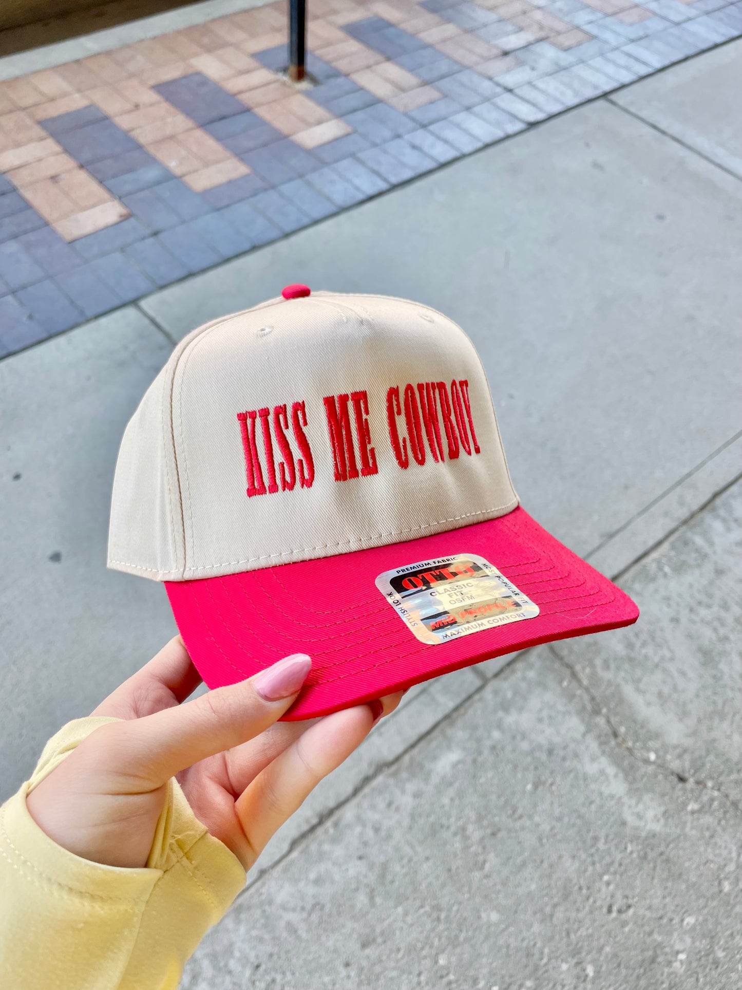Beige and red trucker hat with bold red embroidered text that says "Kiss Me Cowboy," held outdoors on a city sidewalk.
