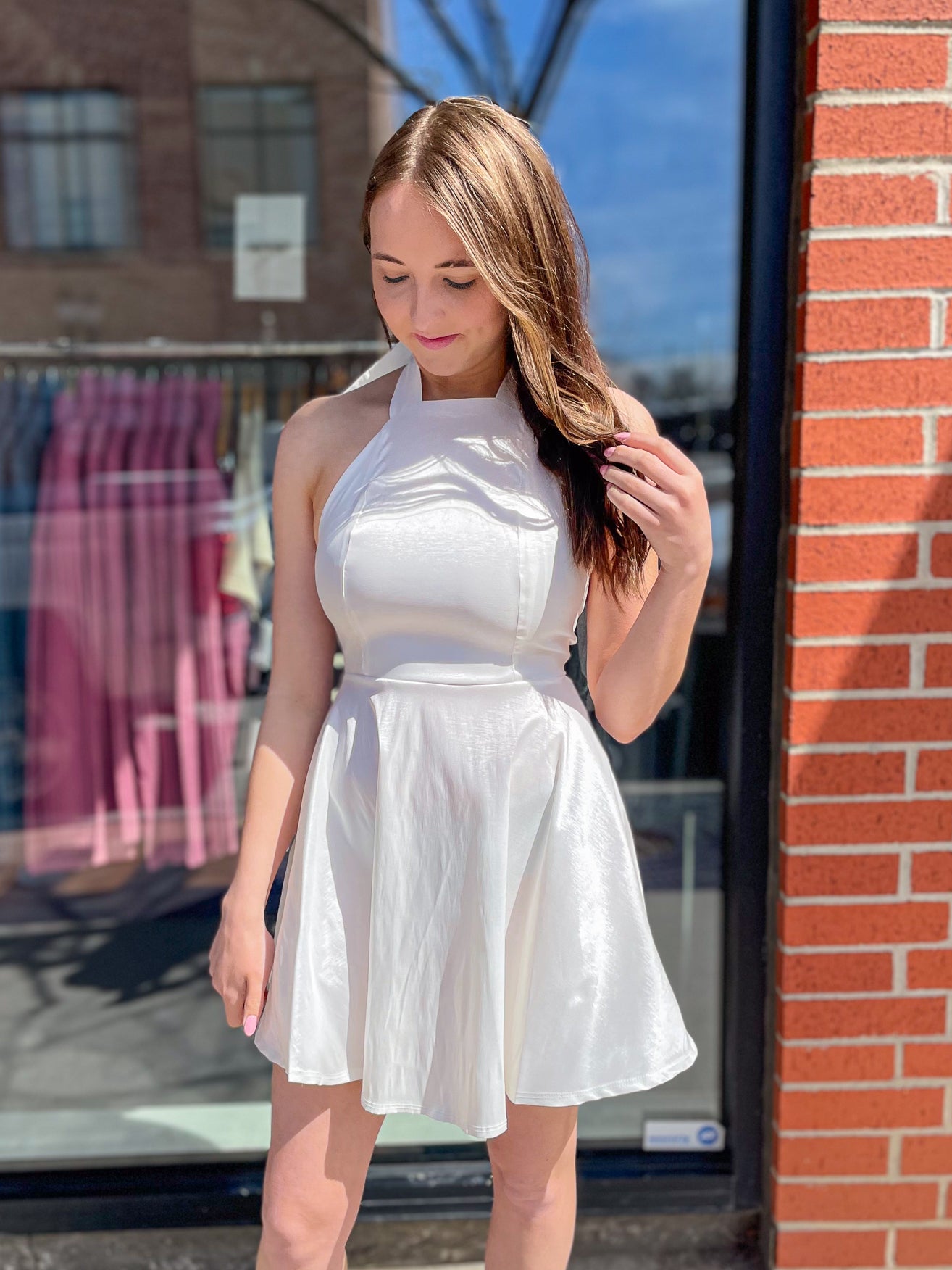 Young woman wearing a white halter dress with a flared skirt, standing outside while looking down and holding her hair.