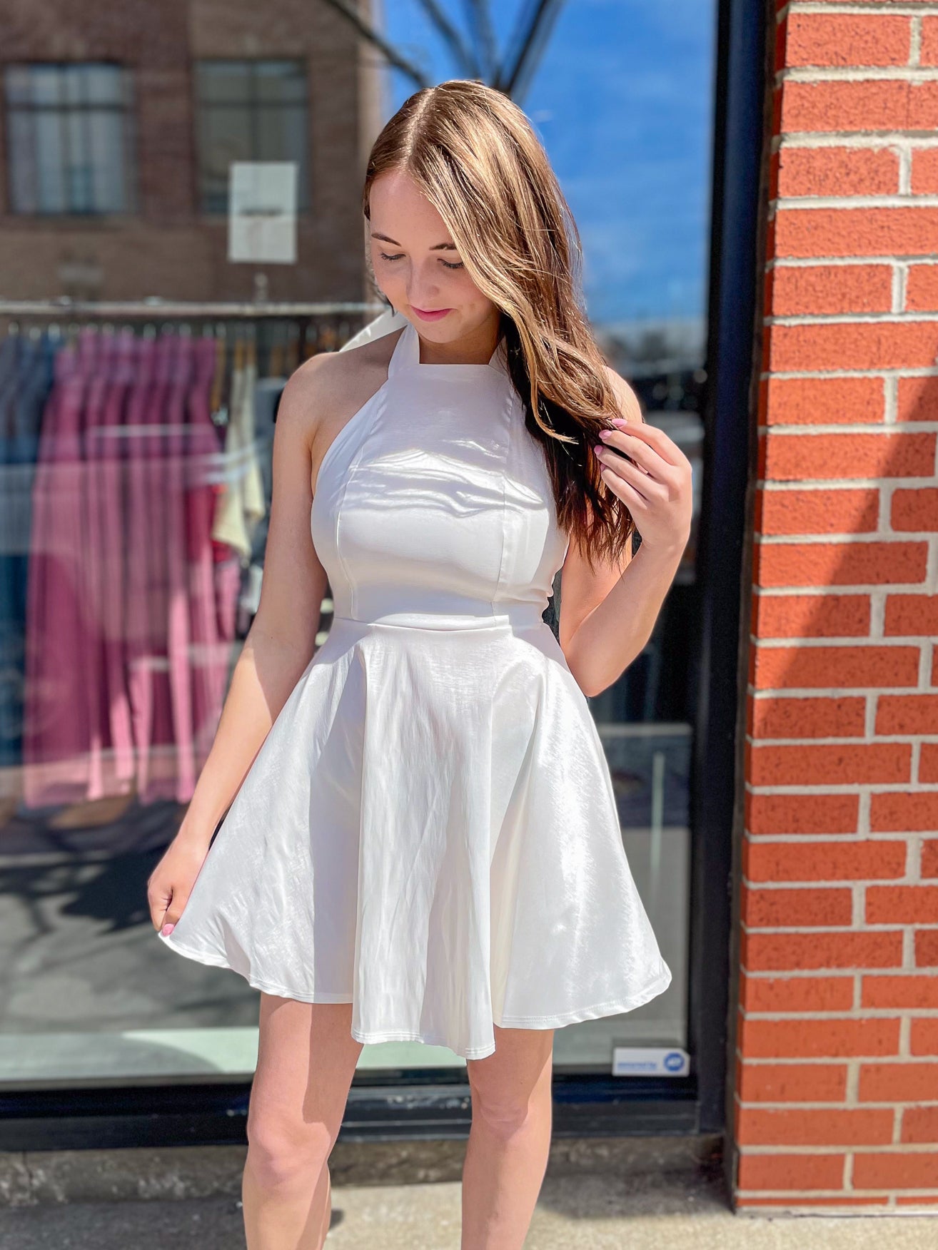 Front view of a woman in a white satin halter dress, holding the hem and looking down with sunlight reflecting off the fabric.