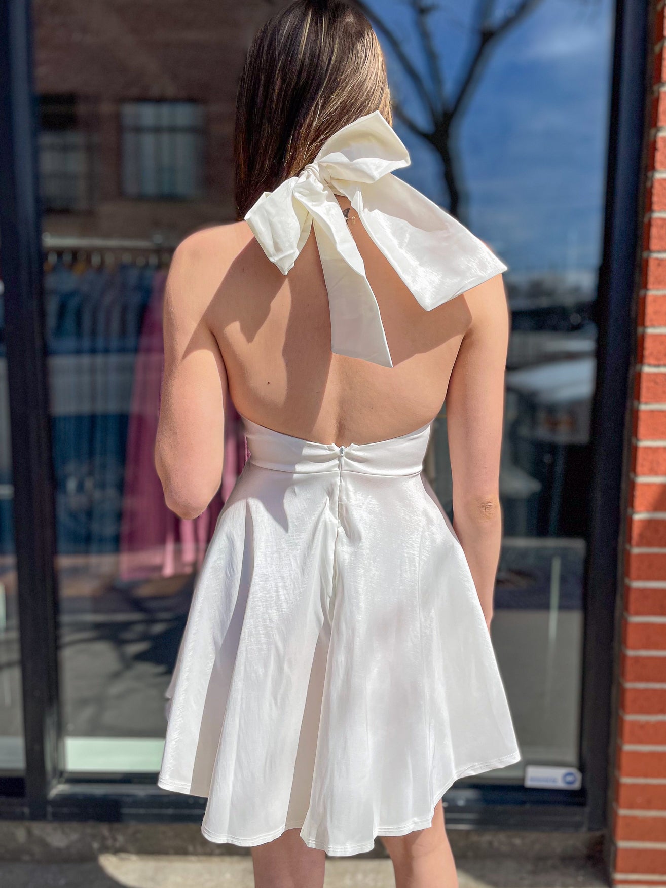 Back view of a white halter dress featuring an oversized bow at the neck and open back detail, worn by a woman standing outside.