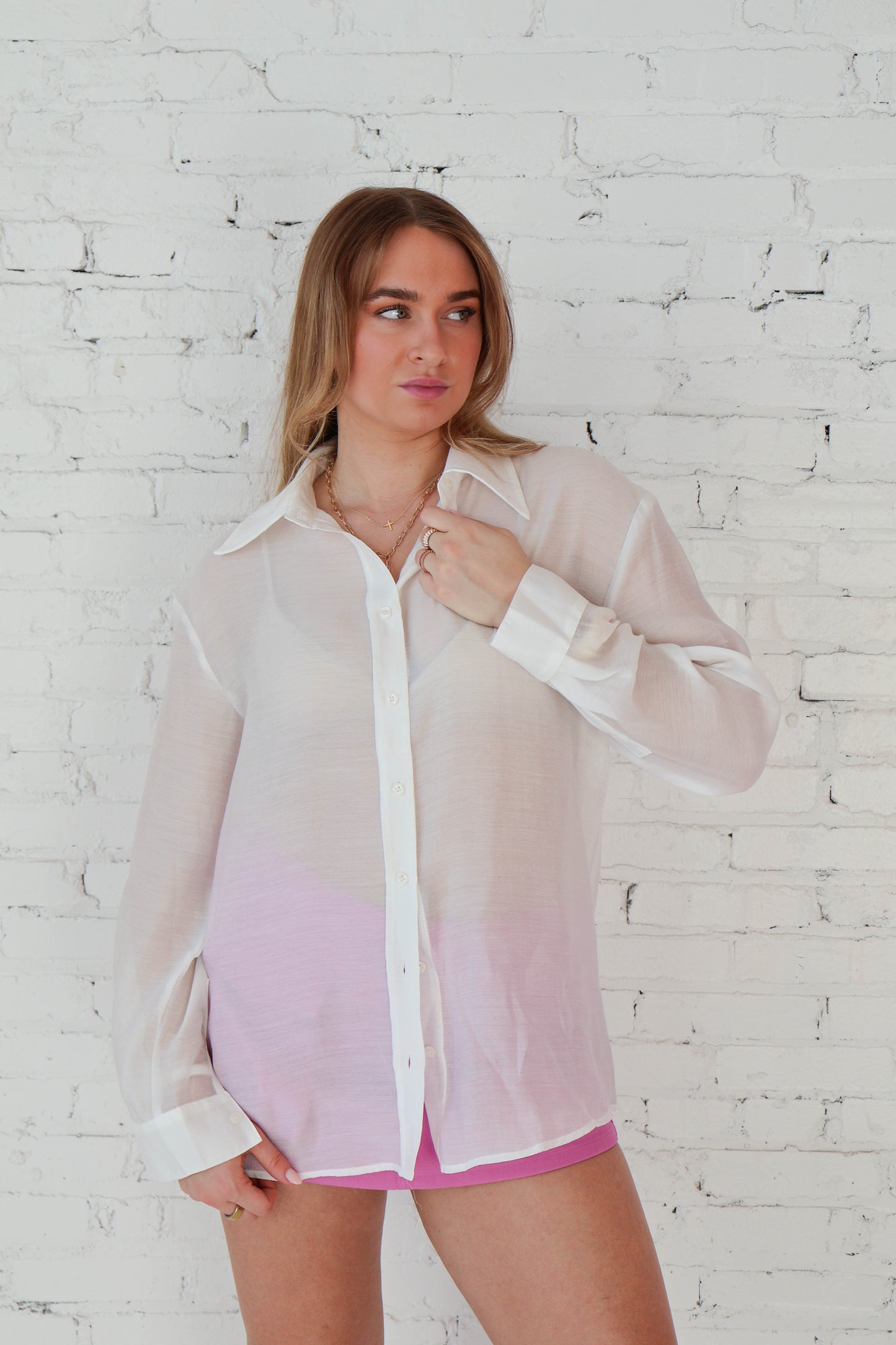 Woman wearing a sheer white blouse with pink trim against a white brick wall.