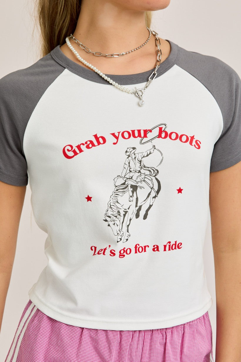 Model wearing a western-style graphic baby tee with grey sleeves and red cowboy text that reads “Grab your boots, let’s go for a ride”