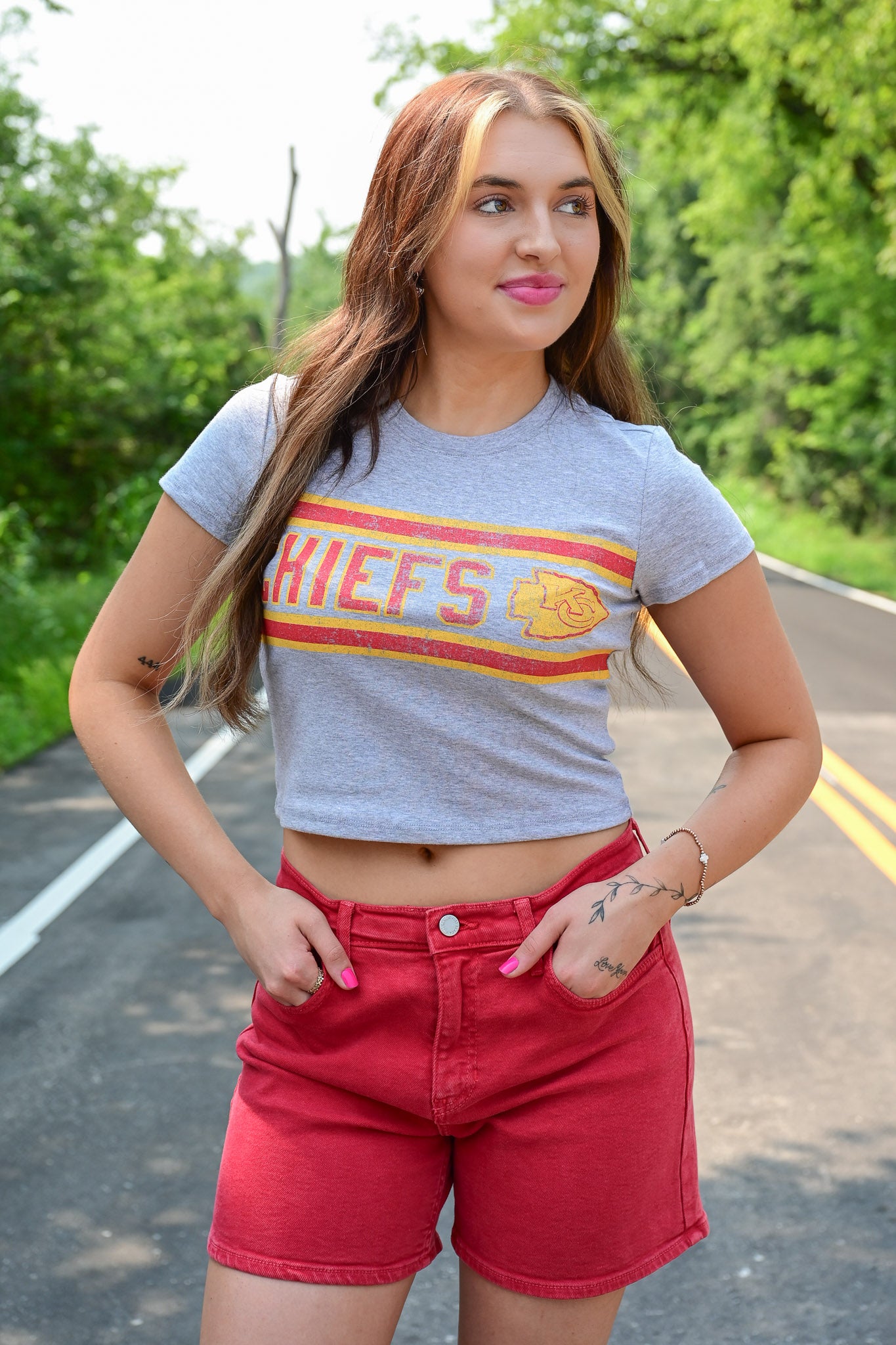 Woman standing on a road wearing a fitted gray crop top with a vintage-style Kansas City Chiefs logo and red high-waisted shorts, with greenery in the background.