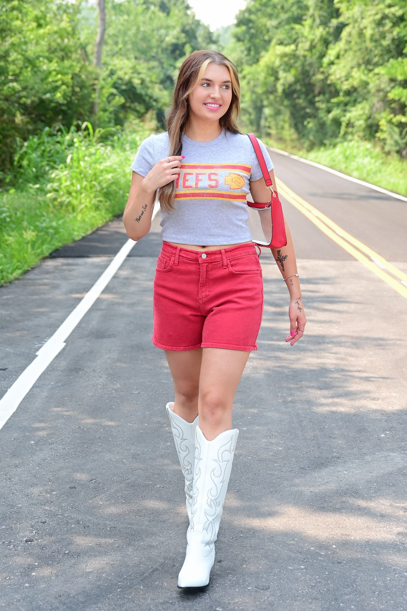 Woman walking down a road in a gray Kansas City Chiefs crop top, red shorts, white embroidered cowboy boots, and a red shoulder bag, smiling with trees lining the road.