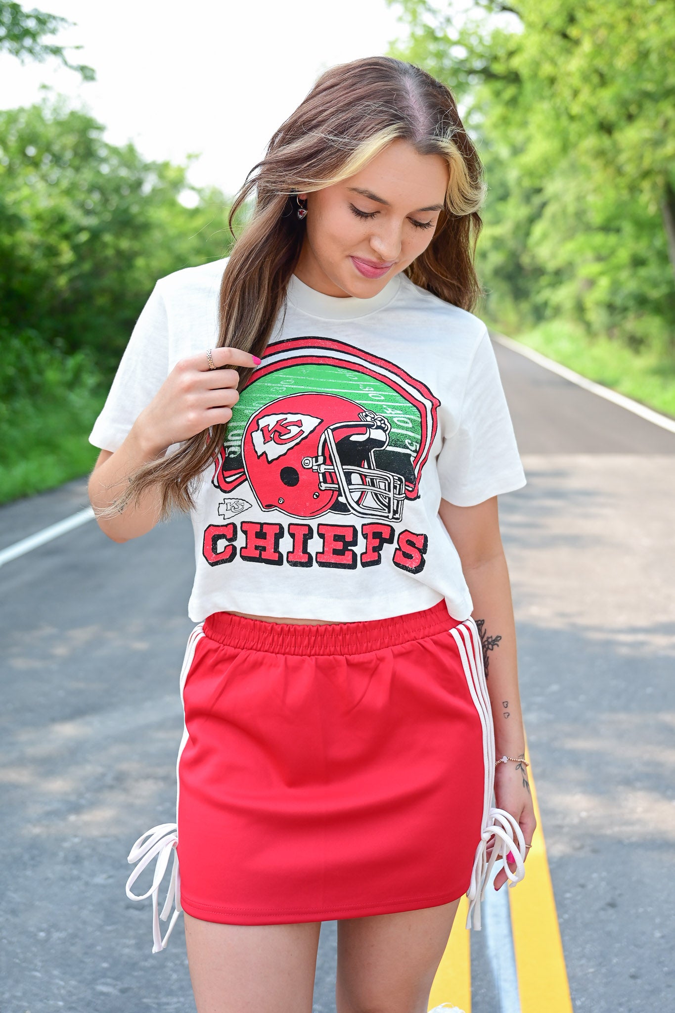 Woman looking down and smiling in a white Chiefs crop top featuring a red helmet and green field graphic, paired with a red skirt with white stripes and side ties, standing on a tree-lined road.