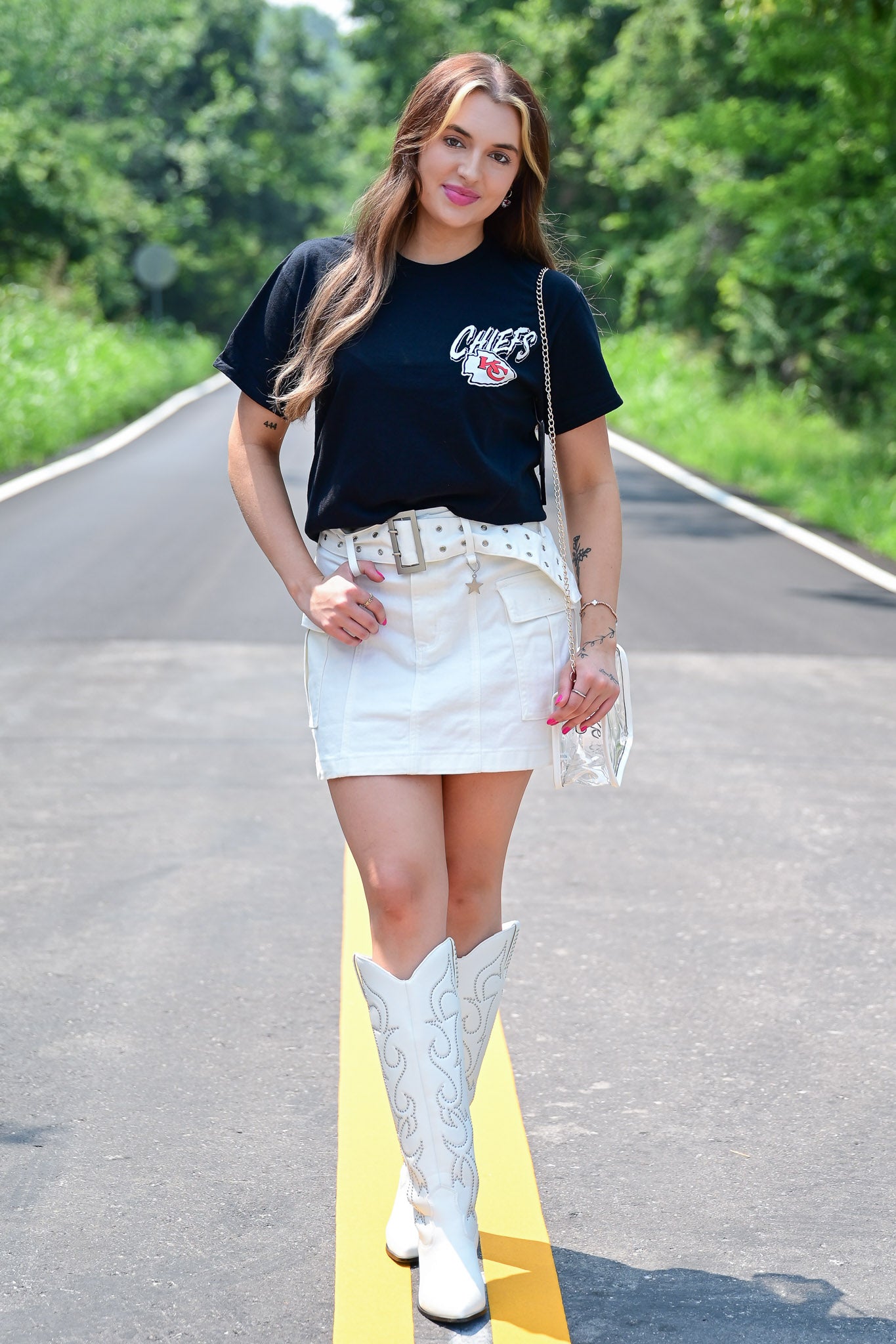 Woman standing confidently on a road wearing a black Kansas City Chiefs t-shirt, white mini skirt with pockets, studded belt, white embroidered cowboy boots, and a clear crossbody purse, with greenery in the background.