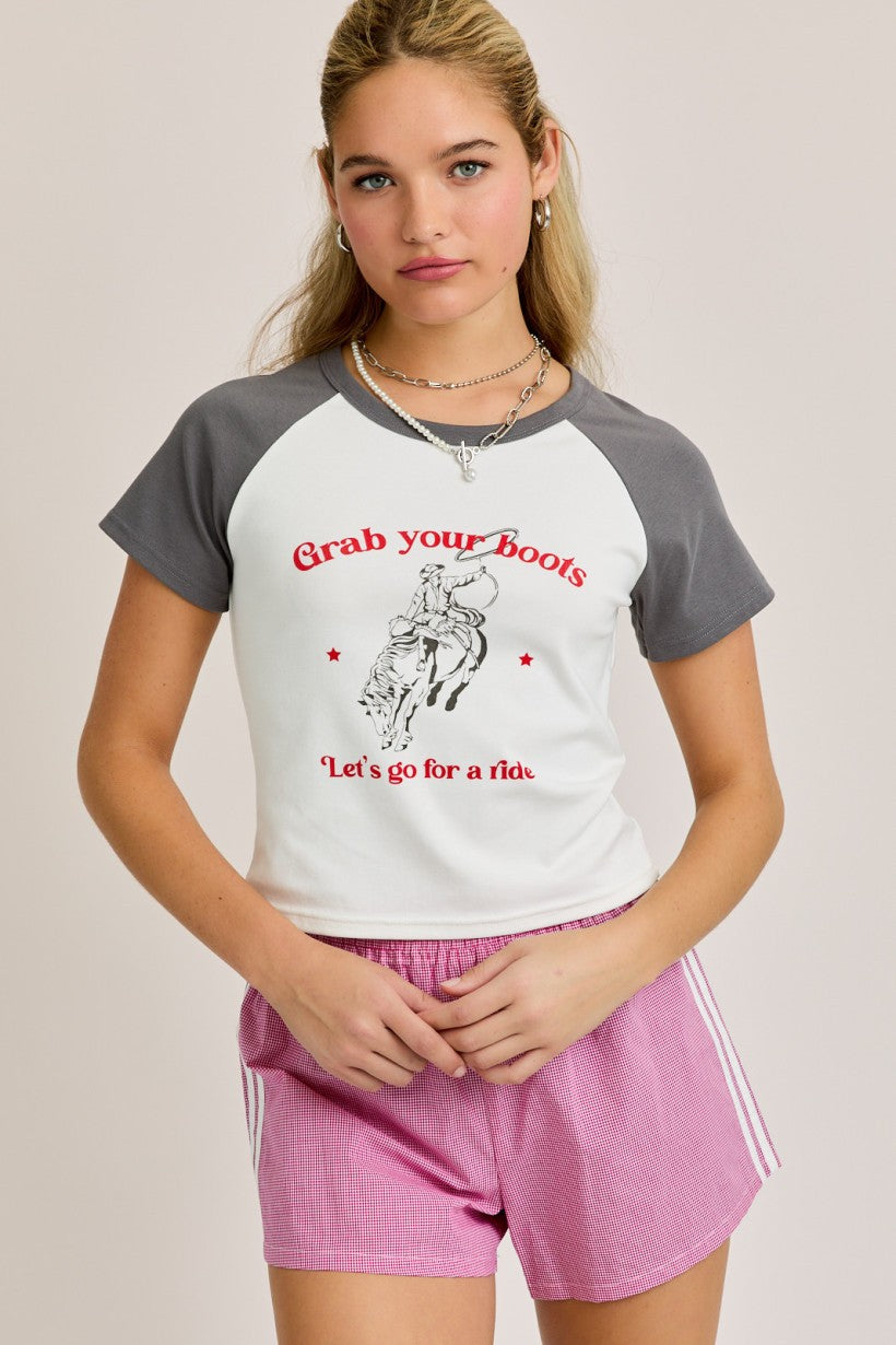 Model wearing a western-style graphic baby tee with grey sleeves and red cowboy text that reads “Grab your boots, let’s go for a ride”