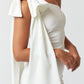 Side view of a woman wearing a white one-shoulder mini dress with a dramatic bow and long draped fabric on the shoulder.