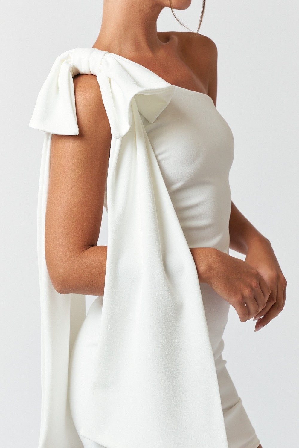 Side view of a woman wearing a white one-shoulder mini dress with a dramatic bow and long draped fabric on the shoulder.