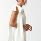 Side view of a woman wearing a white one-shoulder mini dress with a dramatic bow and long draped fabric on the shoulder.