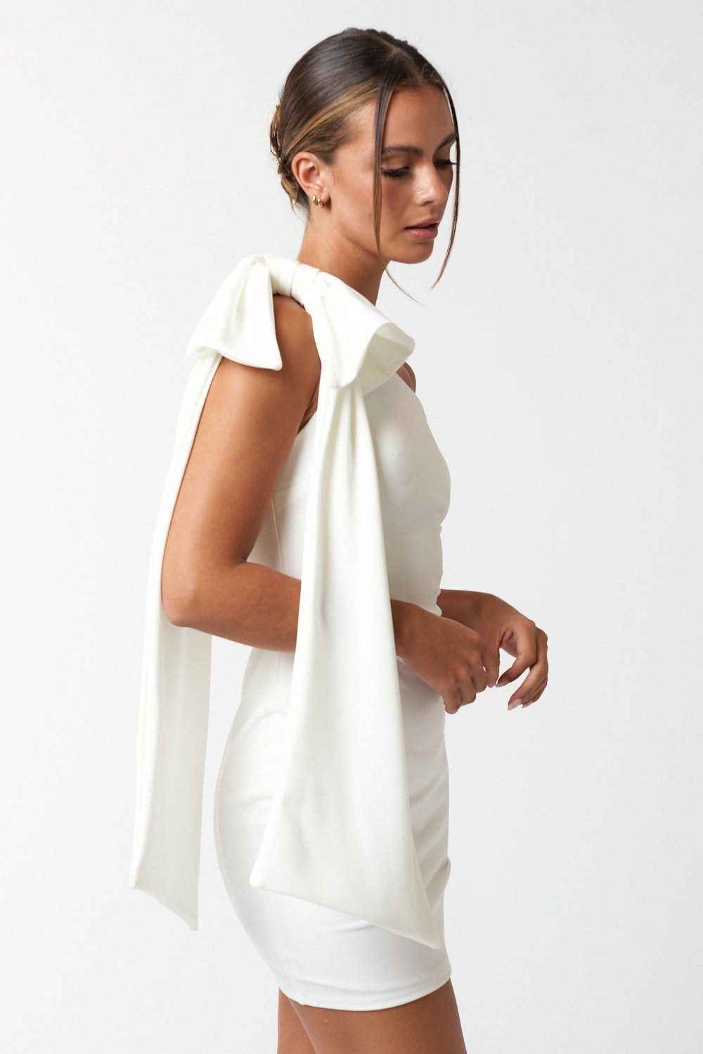 Side view of a woman wearing a white one-shoulder mini dress with a dramatic bow and long draped fabric on the shoulder.