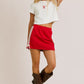 Front view of a red athletic-style mini skirt featuring white side stripes, ruched drawstring ties on both sides, and a stretchy waistband, styled with tall brown buckle boots.