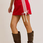 side view of a red athletic-style mini skirt featuring white side stripes, ruched drawstring ties on both sides, and a stretchy waistband, styled with tall brown buckle boots.