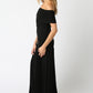 side view of Woman wearing a black off-the-shoulder maxi dress with a fitted bodice and pleated floor-length skirt, standing against a white background.