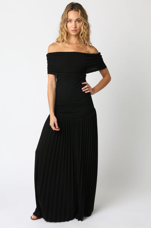 Woman wearing a black off-the-shoulder maxi dress with a fitted bodice and pleated floor-length skirt, standing against a white background.