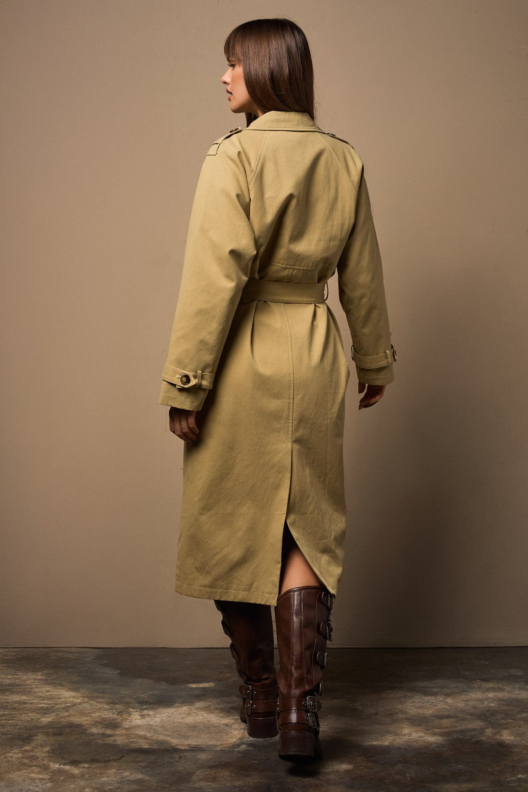 Woman wearing a long beige trench coat and brown boots against a plain background