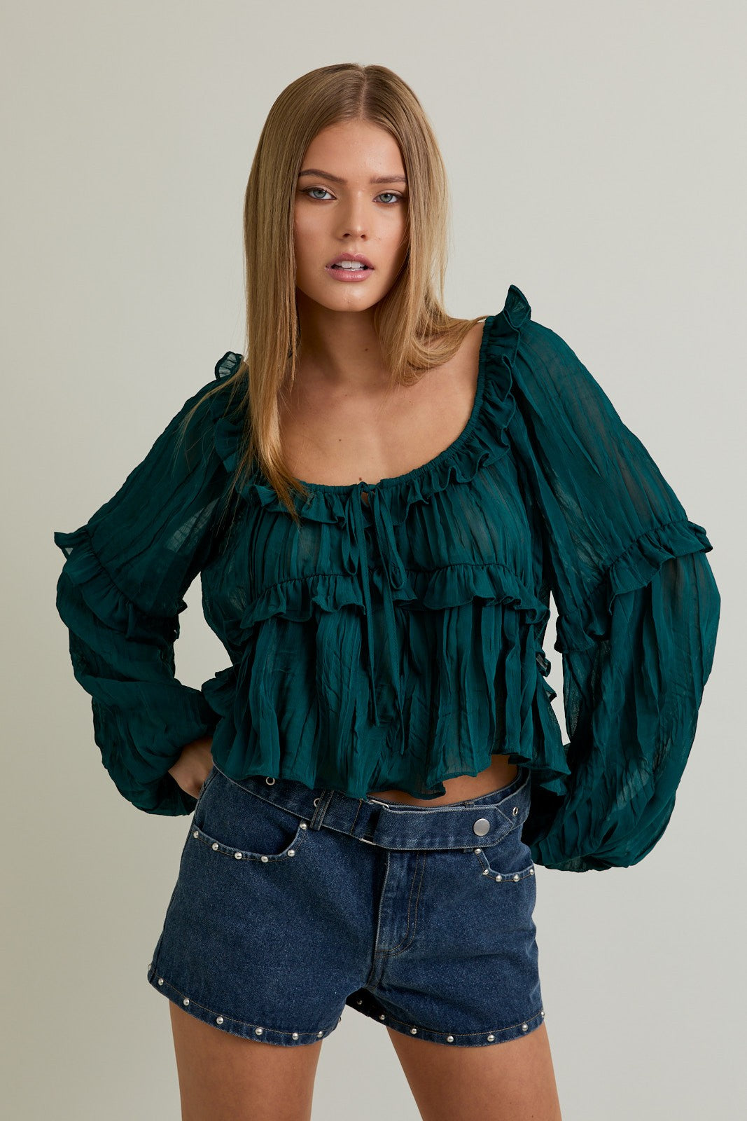 Woman wearing a green ruffled blouse and denim shorts against a neutral background
