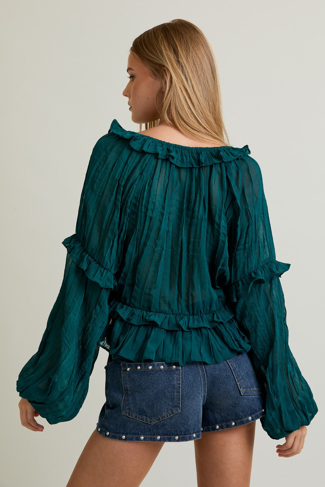 Woman wearing a teal blouse with ruffled details and denim shorts on a plain background