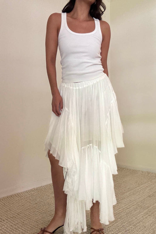 Woman wearing a white tank top and an asymmetrical white ruffle midi skirt, standing in a neutral-toned room with a textured carpet.