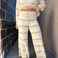 Person wearing a beige and white striped sweater and pants against a brick wall.