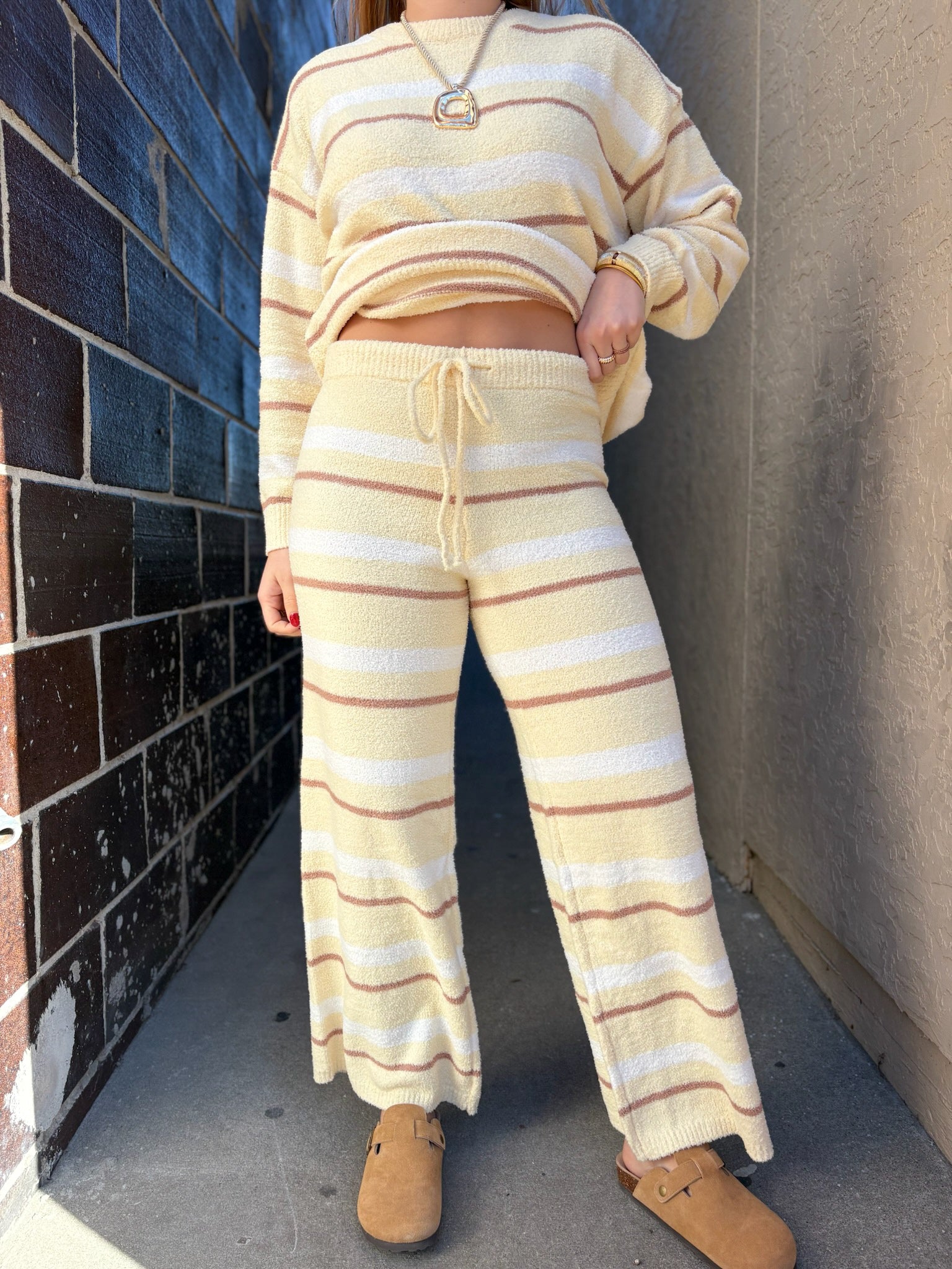 Person wearing a beige and white striped sweater and pants against a brick wall.