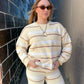 Woman wearing a beige and white striped sweater and pants against a brick wall.