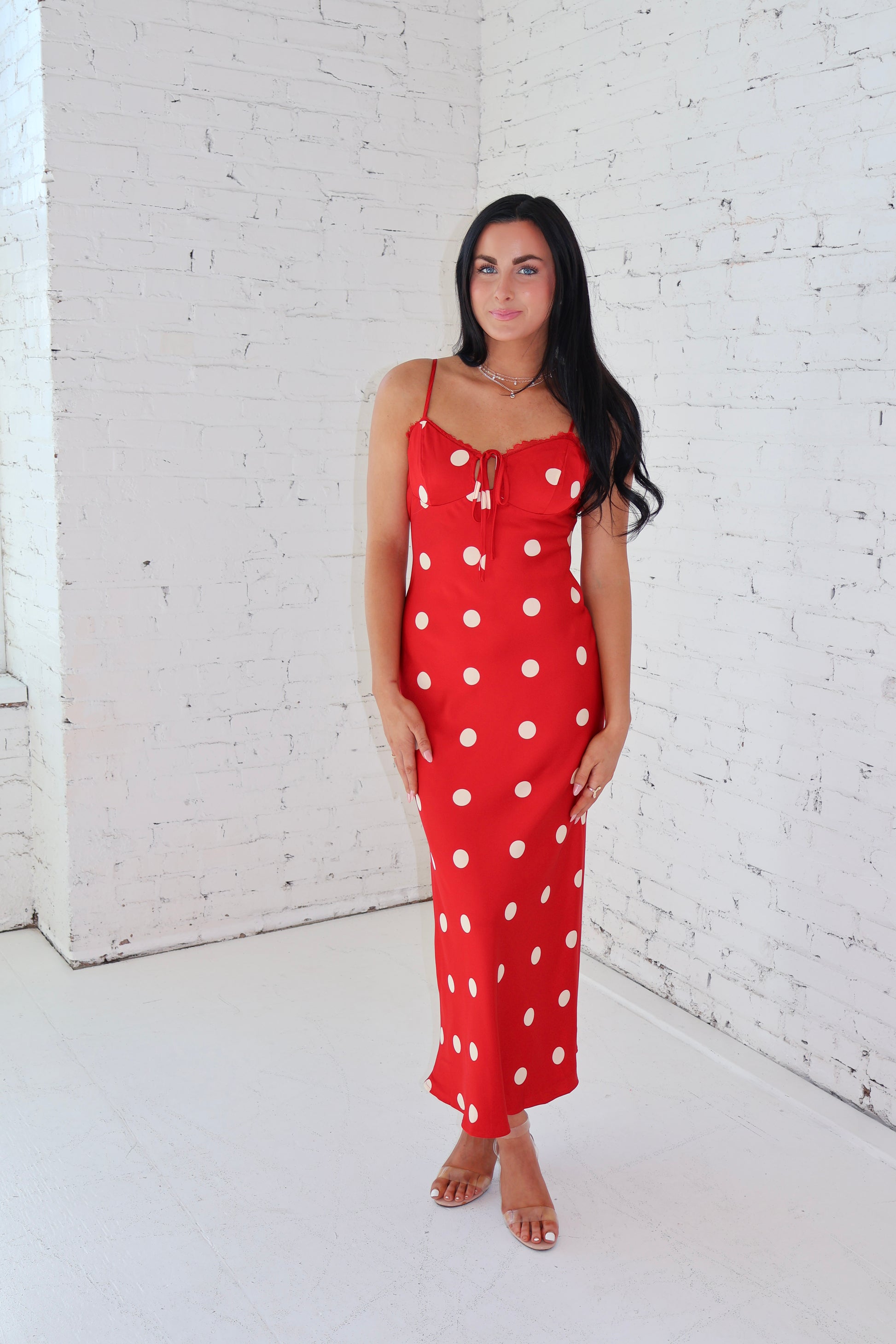 Woman wearing a red polka dot dress against a white brick wall.