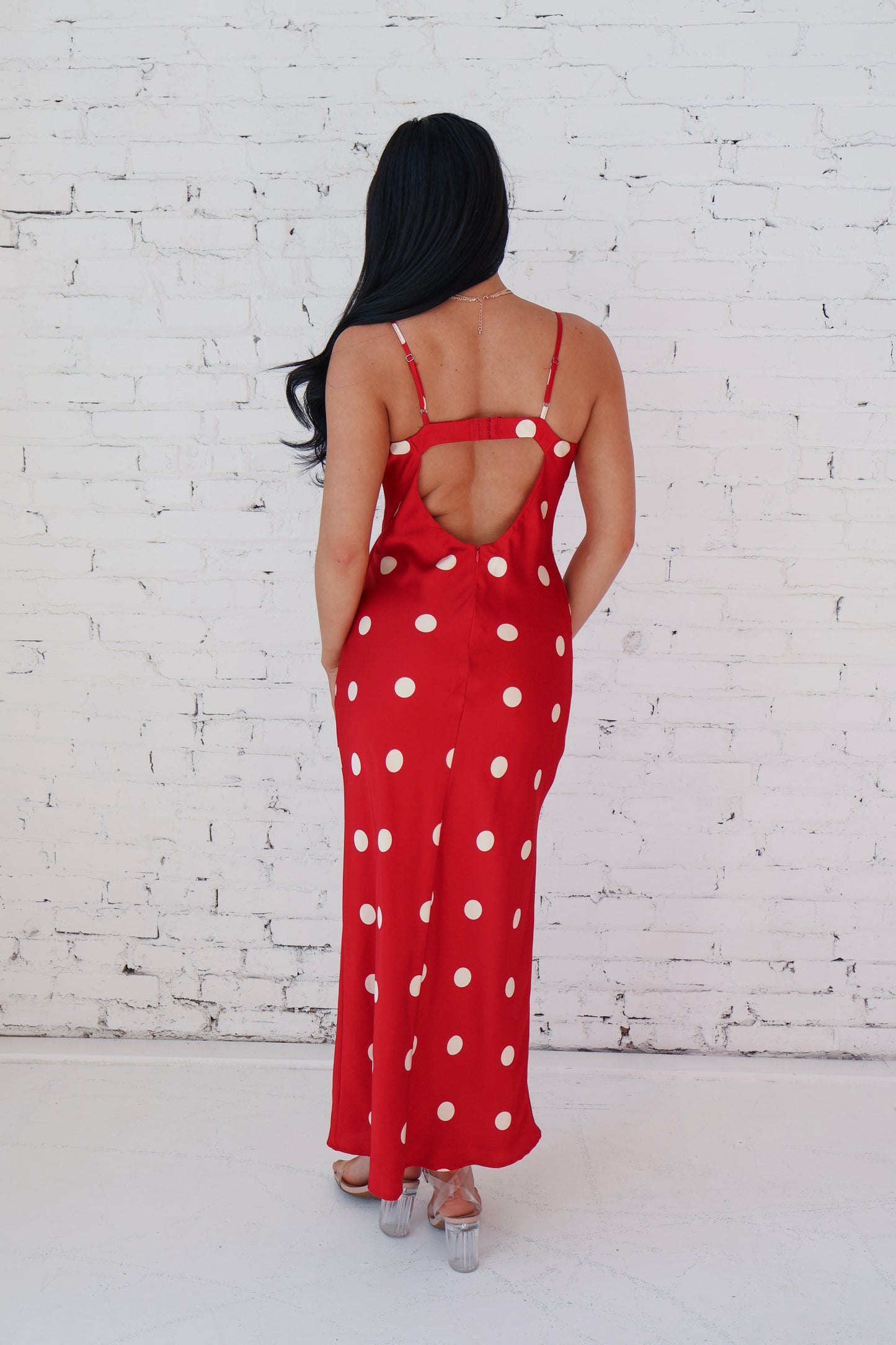 Woman wearing a red polka dot dress with an open back against a white brick wall.