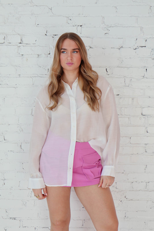 Woman wearing a sheer white shirt over a pink skirt against a white brick wall.