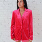 Woman wearing a bright pink blazer over a checkered dress against a white brick wall.