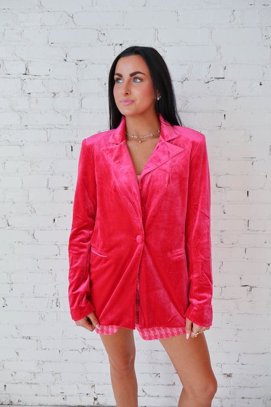 Woman wearing a bright pink blazer over a checkered dress against a white brick wall.