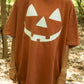 Person wearing a brown t-shirt with a white jack-o'-lantern face design in a forest setting