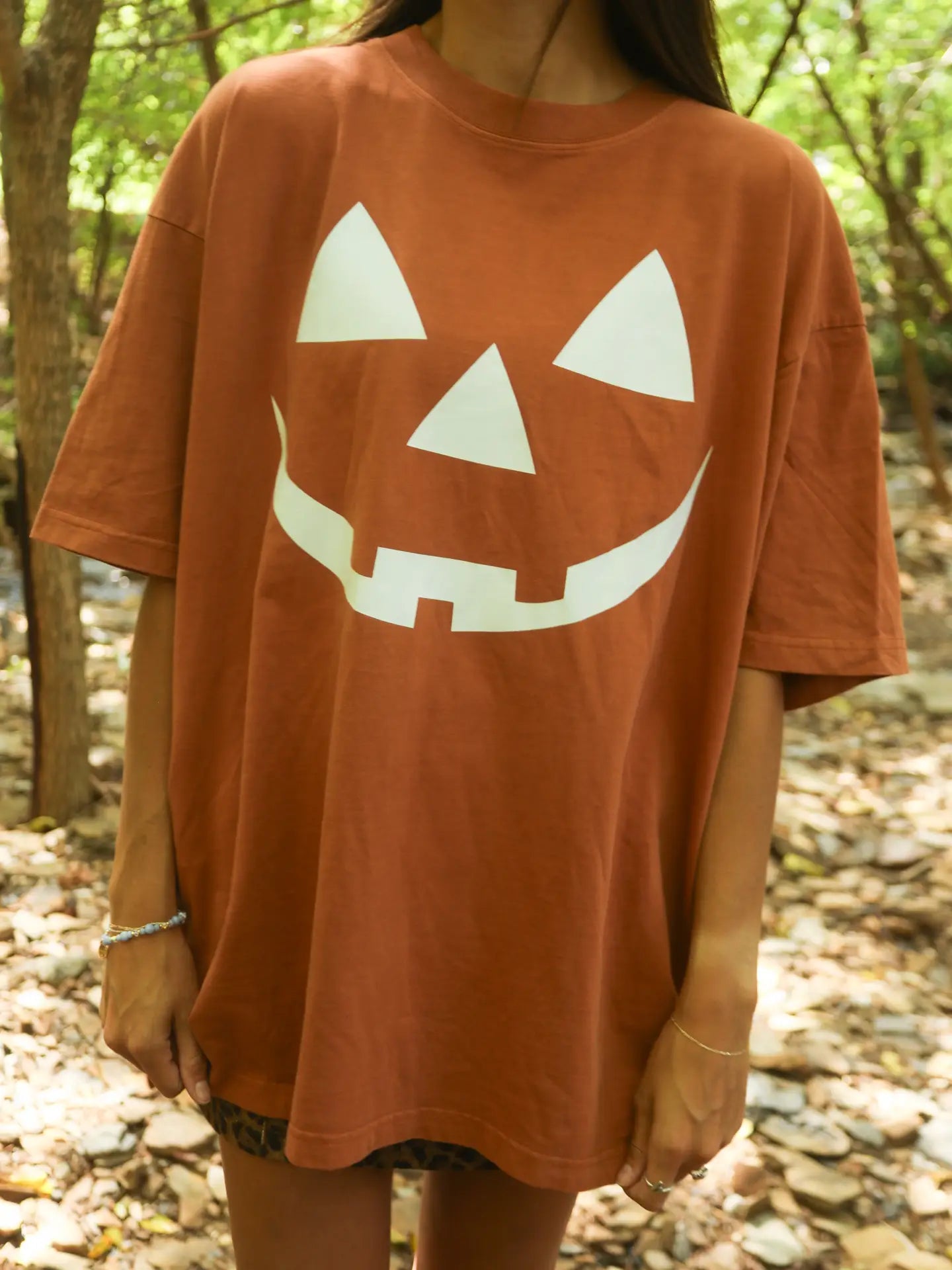 Person wearing a brown t-shirt with a white jack-o'-lantern face design in a forest setting